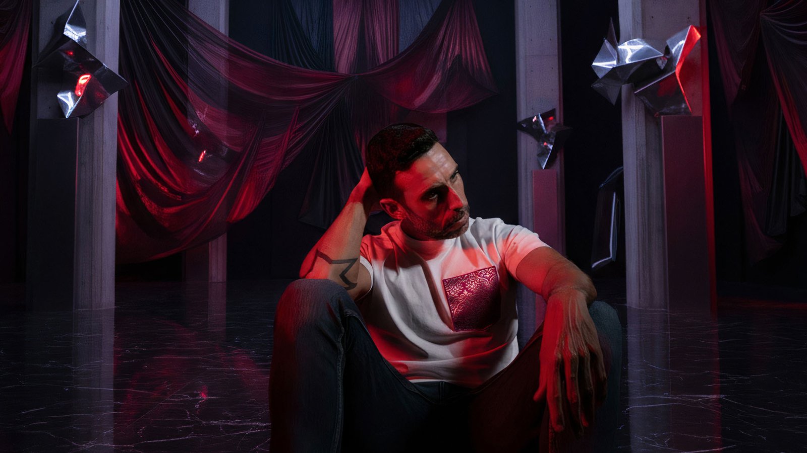 Hombre sentado en un sillón de estilo trono con la camiseta blanca faaia, en un ambiente dramático con telas y luces rojas