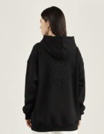 Mujer joven de espaldas con sudadera negra oversize marca Faaia que lleva un bordado ornamental en la espalda y tela damasco en la capucha.
