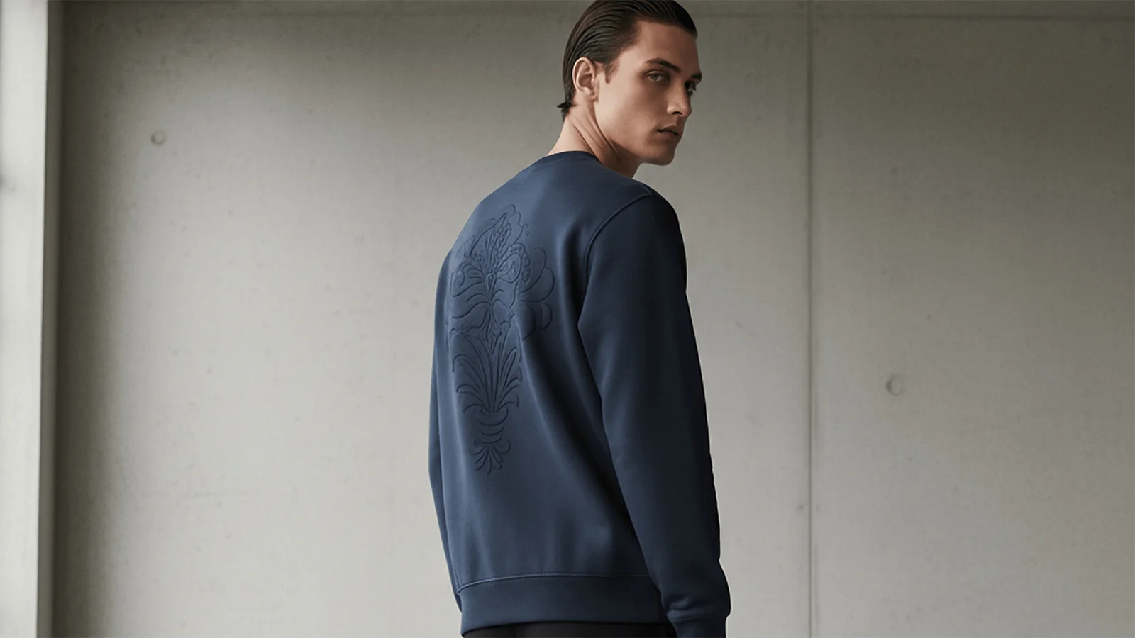 Hombre joven con pelo castaño mirando hacia atrás con una sudadera azul marino Faaia con bordado de damasco valenciano al tono.