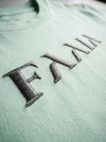 Primer plano de el logo de Faaia bordado en hilo plata sobre camiseta color verde agua.