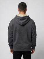 hombre joven posando de espaldas con una sudadera color gris con la capucha forrada con un damasco blanco y dorado y con un bordado ornamental en la espalda.